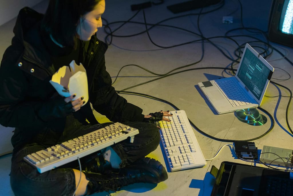 A young woman in a dark room surrounded by computers and cables, eating and typing on keyboards.