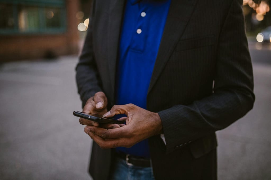 A man in a black suit using a smartphone while standing outdoors during the day.