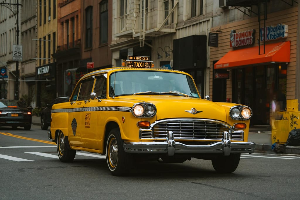 Vintage yellow taxi cab driving through bustling New York City street with iconic buildings.