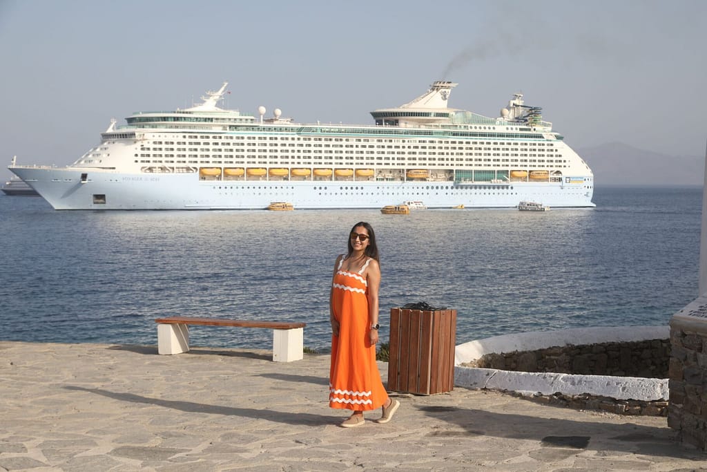 A woman in an orange dress smiles on the Mykonos waterfront with a cruise ship in the background.