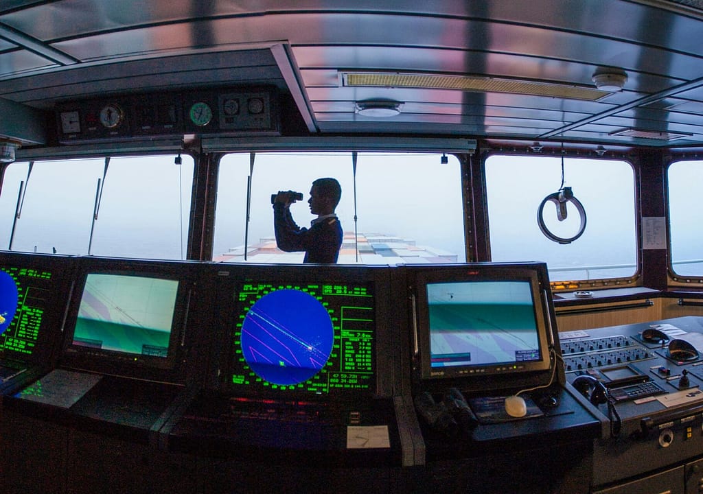 Silhouette of a navigator using binoculars on a cargo ship's bridge, surrounded by navigation equipment.
