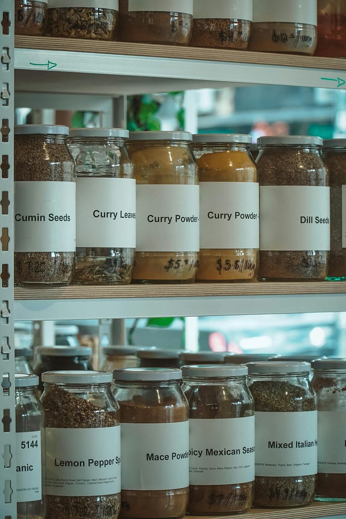 Close-up shot of various spices in labeled glass jars on wooden shelves in a store.