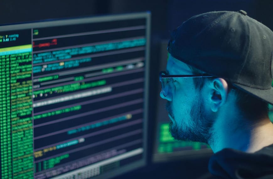 A cybersecurity expert inspecting lines of code on multiple monitors in a dimly lit office.