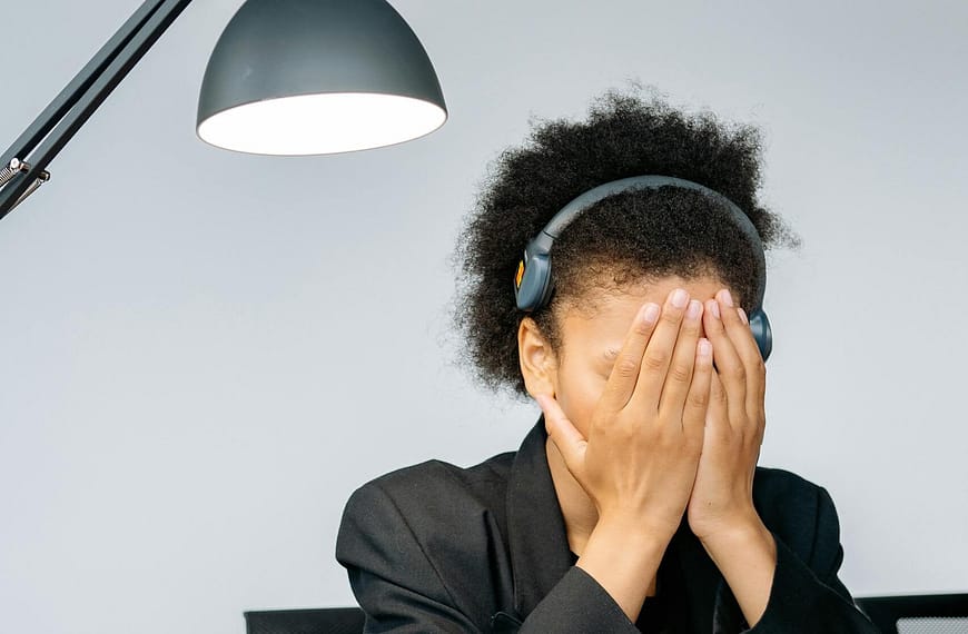 A tired call center employee covering her face with hands in frustration, wearing a headset in an office.