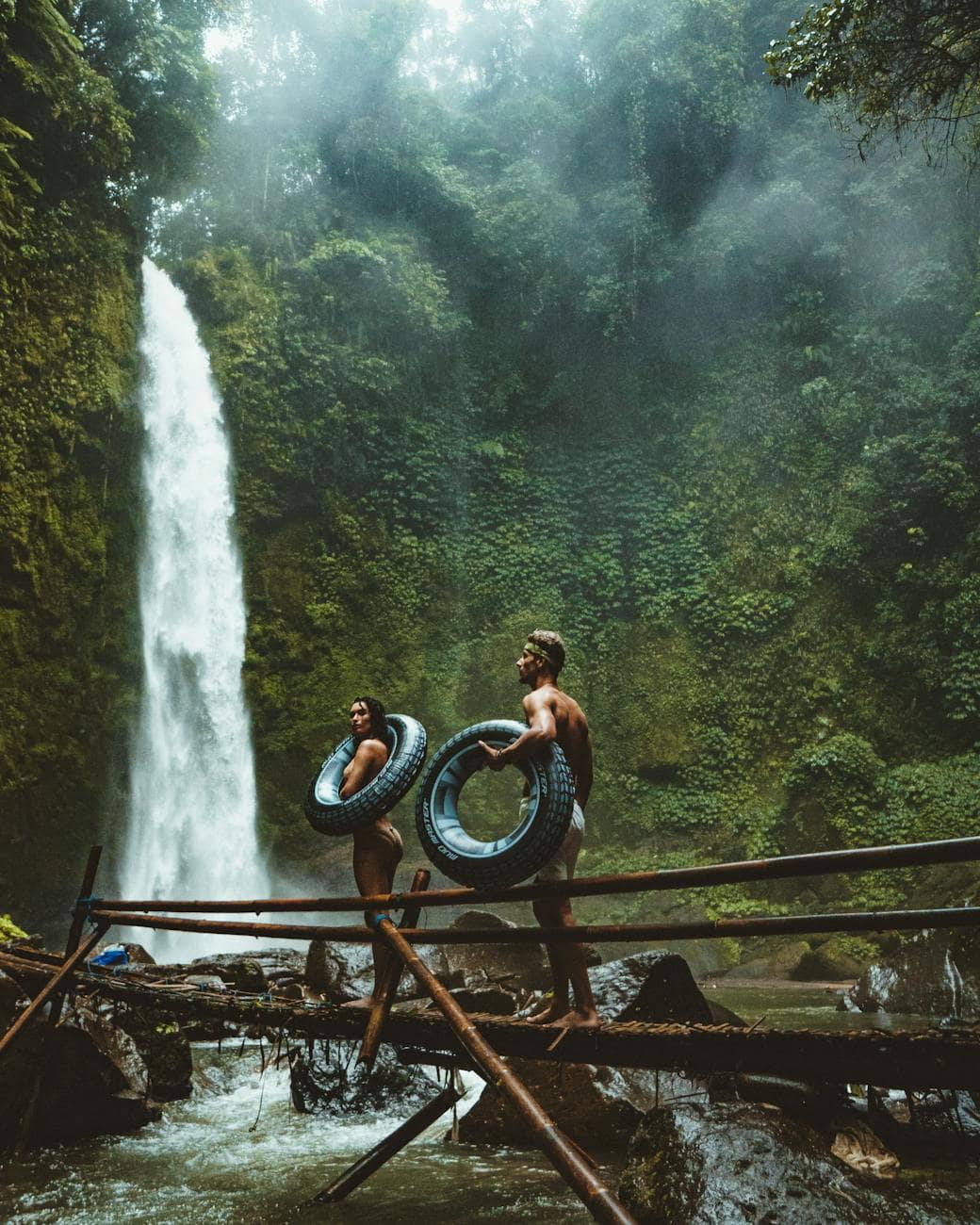 two person carrying black inflatable pool float on brown wooden bridge near waterfalls/ Photo by Oliver Sjöström on Pexels.com